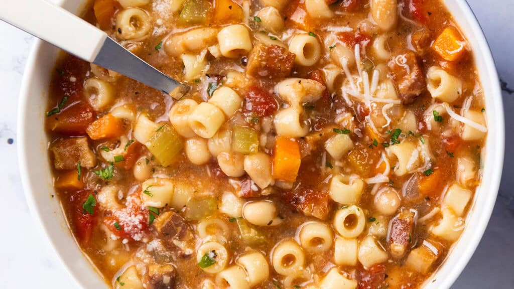 A close-up of a bowl of Pasta Fagioli alla Veneta soup, showcasing ditalini pasta, diced carrots, celery, and tomatoes. The dish is elegantly garnished with shredded Parmesan and parsley, with a spoon resting invitingly in the bowl.