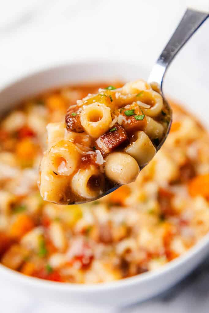 A close-up of a spoonful of traditional Pasta Fagioli alla Veneta, highlighting small pasta tubes, kidney beans, and carrots garnished with herbs. In the background, a bowl brims with this hearty Veneta soup.