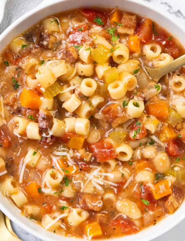 A bowl of Pasta Fagioli alla Veneta soup filled with pasta, diced tomatoes, carrots, beans, and celery, garnished with grated cheese and parsley. This classic dish of Italian cuisine boasts a hearty, rustic appearance with a variety of vibrant colors.