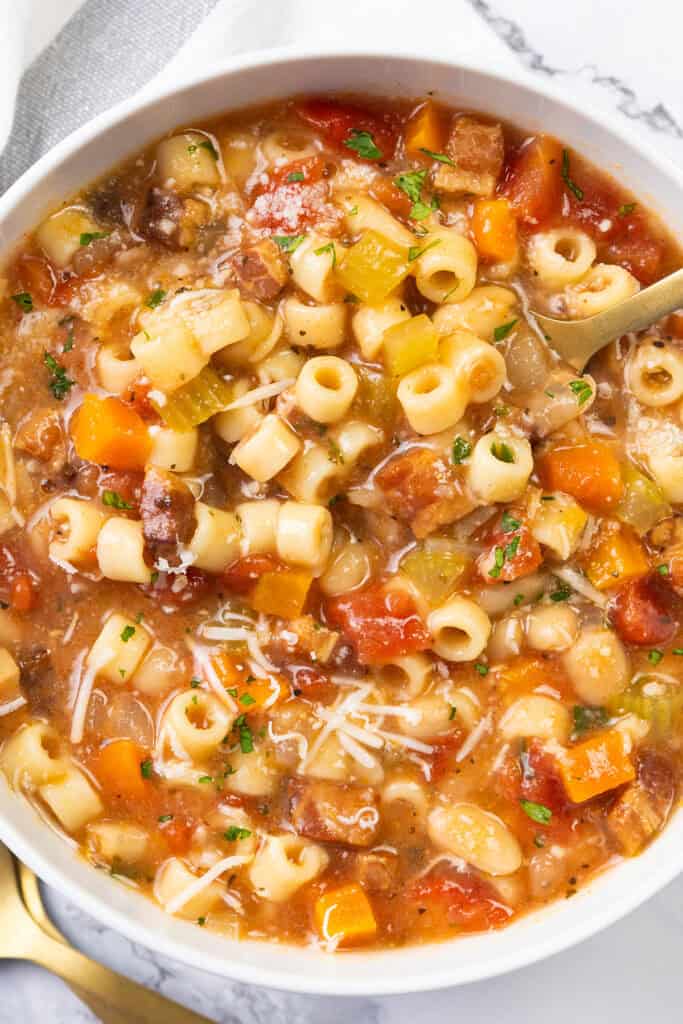 A bowl of Pasta Fagioli alla Veneta soup filled with pasta, diced tomatoes, carrots, beans, and celery, garnished with grated cheese and parsley. This classic dish of Italian cuisine boasts a hearty, rustic appearance with a variety of vibrant colors.