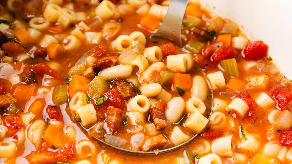 A ladle scoops minestrone soup from a single pot, featuring ditalini pasta, white beans, diced tomatoes, carrots, celery, and parsley in a rich tomato-based broth. Perfect for those who love effortless one-pot recipes.