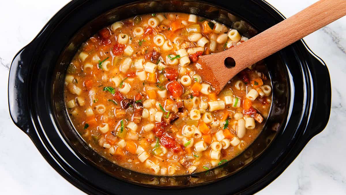 A black crockpot filled with vegetable pasta soup, featuring small pasta shapes, diced tomatoes, onions, and herbs, with a wooden spoon resting inside. The soup appears hearty and colorful, placed on a light marble surface.