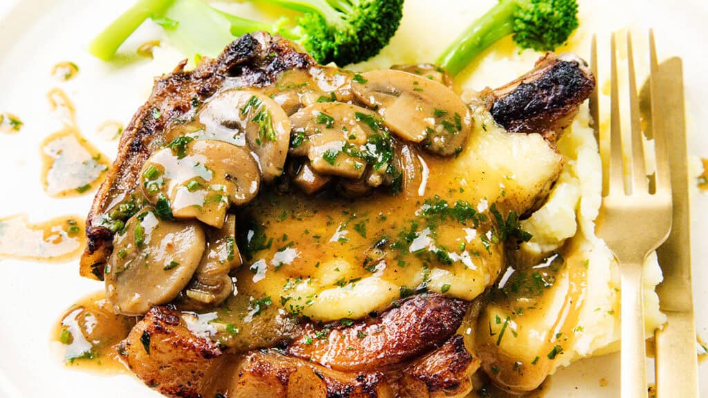 A plate with a pork chop crowned with sliced mushrooms and gravy, garnished with herbs. Served alongside creamy mashed potatoes and broccoli, this family favorite is sure to be one of those incredible recipes you'll want to make again. A fork and knife are placed beside the steak.