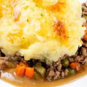 Close-up of a plate of cottage pie, showcasing a golden-brown mashed potato topping over a savory filling of ground meat, peas, carrots, and rich gravy. The dish appears hot and freshly baked.