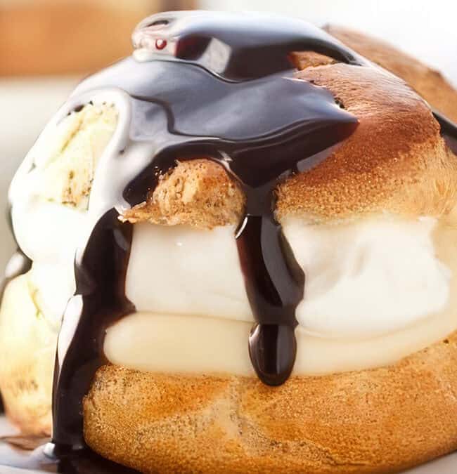 A cream puff topped with a generous drizzle of glossy chocolate sauce, sitting on a white plate. The puff is filled with creamy white filling, contrasting with the golden brown pastry. A spoon rests beside it.