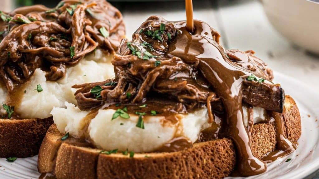 Slices of bread topped with mashed potatoes and shredded beef, covered in brown gravy. Finely chopped herbs garnish the dish, and more gravy is being poured over the top.