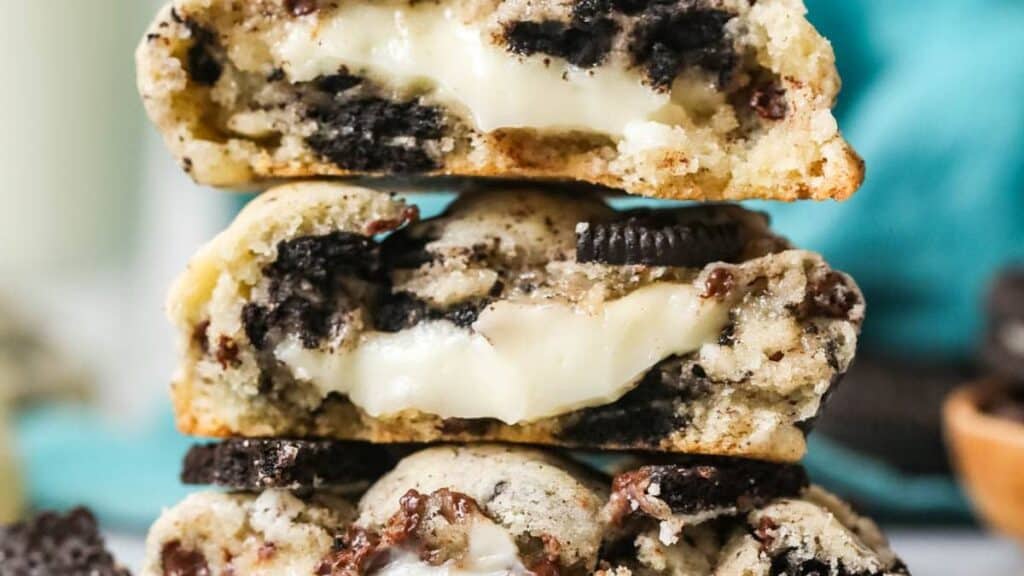 Three cookies stacked on top of each other, revealing a gooey, creamy center. The cookies have a textured surface with visible chocolate cookie pieces throughout. They are set against a blurred background with hints of blue and more cookies.