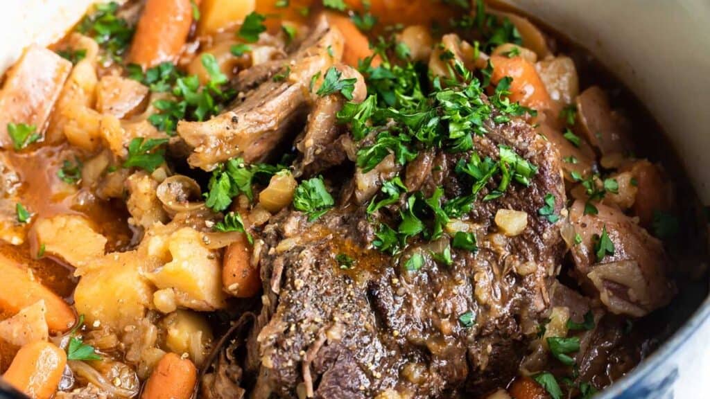 A pot filled with a classic, old-fashioned pot roast surrounded by carrots, potatoes, and onions. The dish is garnished with chopped parsley, and there is a rich, savory sauce visible in the pot.