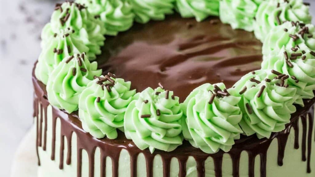 A chocolate cake with mint green frosting and chocolate drizzle spills over the sides. Swirls of mint green frosting are topped with chocolate sprinkles around the edge.