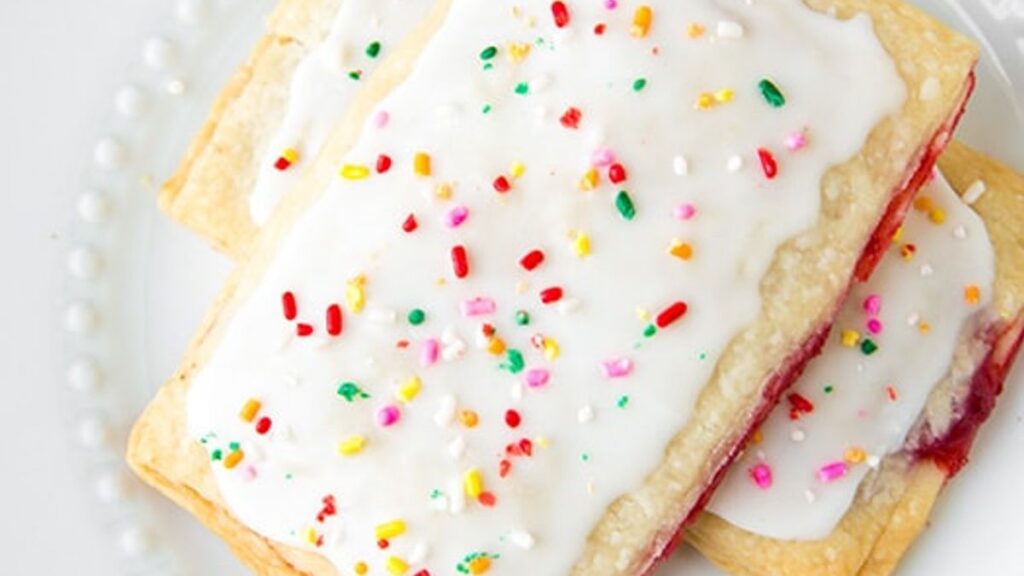 Two frosted pastries with colorful sprinkles are stacked on a white plate. The pastries have a fruit filling and a flaky crust.