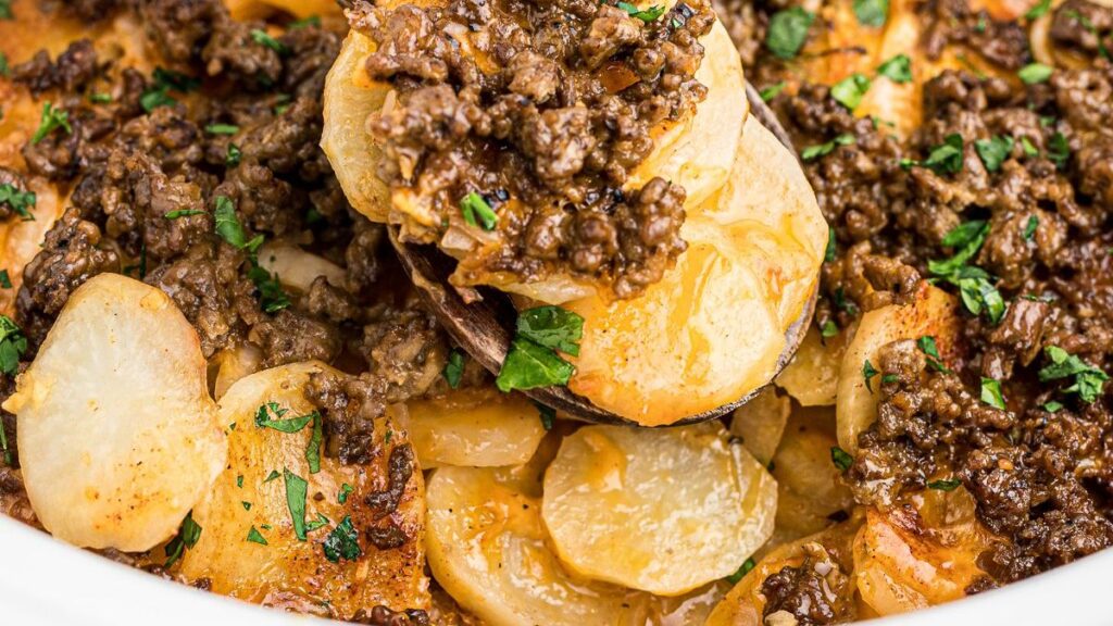 A close-up of a savory dish made in a slow cooker, featuring sliced potatoes layered with ground meat, topped with fresh chopped parsley. The dish is cooked to a golden brown, with some pieces slightly crispy and garnished with herbs.