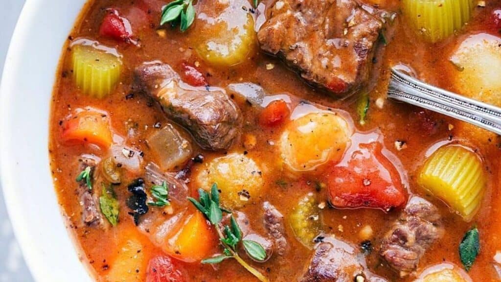 A bowl of delicious beef stew with chunks of beef, potatoes, carrots, celery, and tomatoes in a rich broth. Perfect for slow dinners, it’s garnished with fresh herbs and has a spoon resting in the stew.