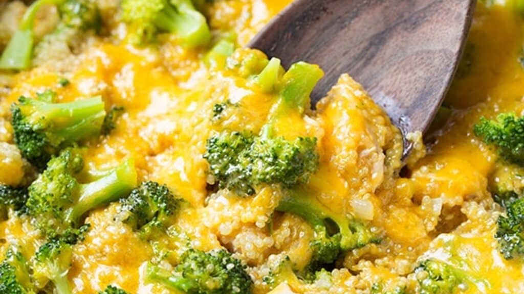 Close-up of a wooden spoon scooping creamy broccoli and quinoa casserole topped with melted cheddar cheese, perfect for an easy one-pot dinner. The dish is vibrant, with the green broccoli contrasting the yellow cheese, creating a hearty and inviting appearance.