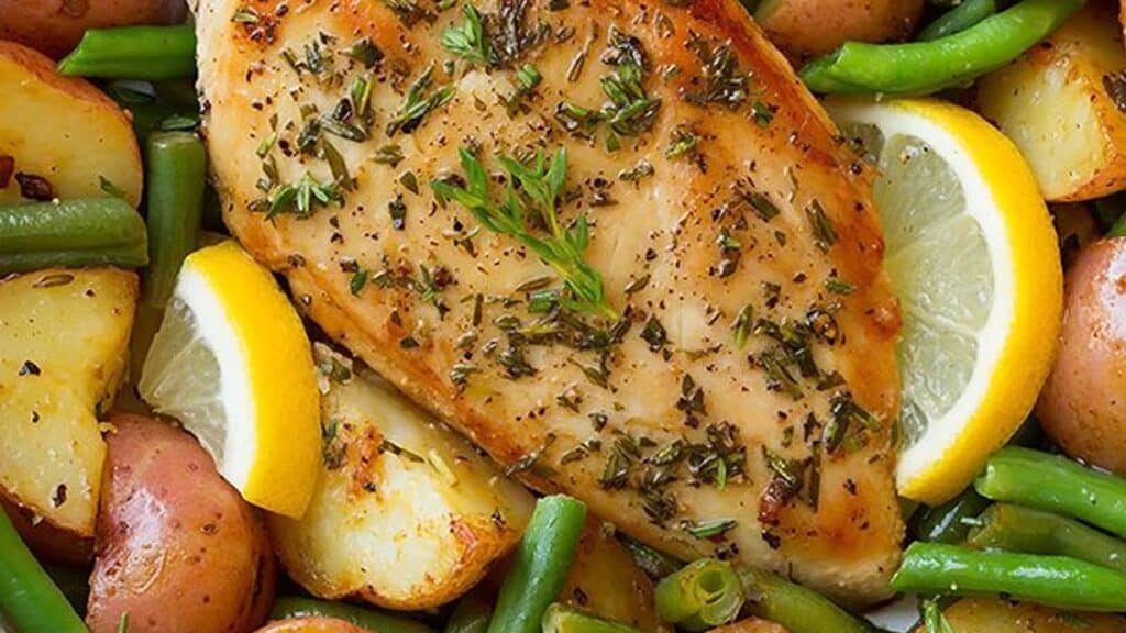 Grilled herb-seasoned chicken breast rests on a bed of roasted red potatoes and green beans with lemon wedges. This flavor-packed dinner is garnished with fresh herbs, creating a vibrant and appetizing presentation that's both delicious and time-saving.