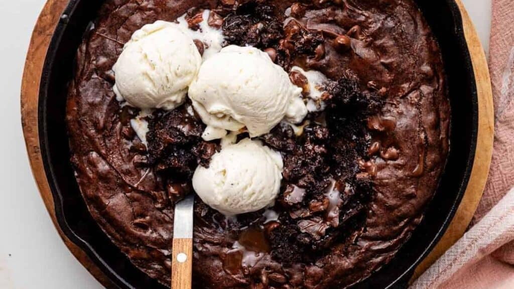 A skillet brownie topped with three scoops of vanilla ice cream. The dark chocolate brownie is served in a round, black skillet on a wooden board, with a knife resting on the edge. A pink cloth is partially visible on the side.