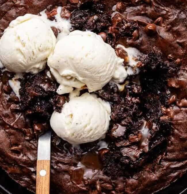 A skillet brownie topped with three scoops of vanilla ice cream. The dark chocolate brownie is served in a round, black skillet on a wooden board, with a knife resting on the edge. A pink cloth is partially visible on the side.