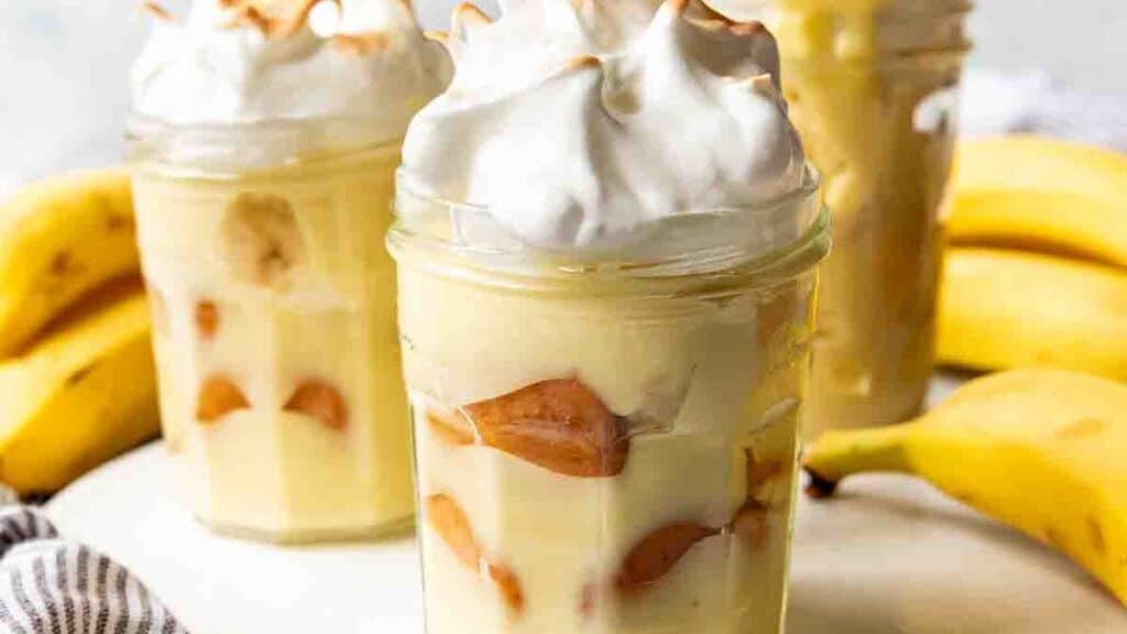 Three mason jars filled with banana pudding, topped with meringue, are surrounded by ripe bananas. The jars include layers of pudding and wafers, and the meringue is lightly toasted. A striped cloth napkin is partially visible.