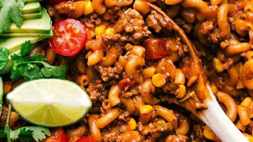 A close-up of a flavorful pasta dish featuring macaroni, ground beef, corn, and sliced cherry tomatoes. Garnished with fresh cilantro, sliced avocado, and a lime wedge, with a wooden spoon stirring the mixture.