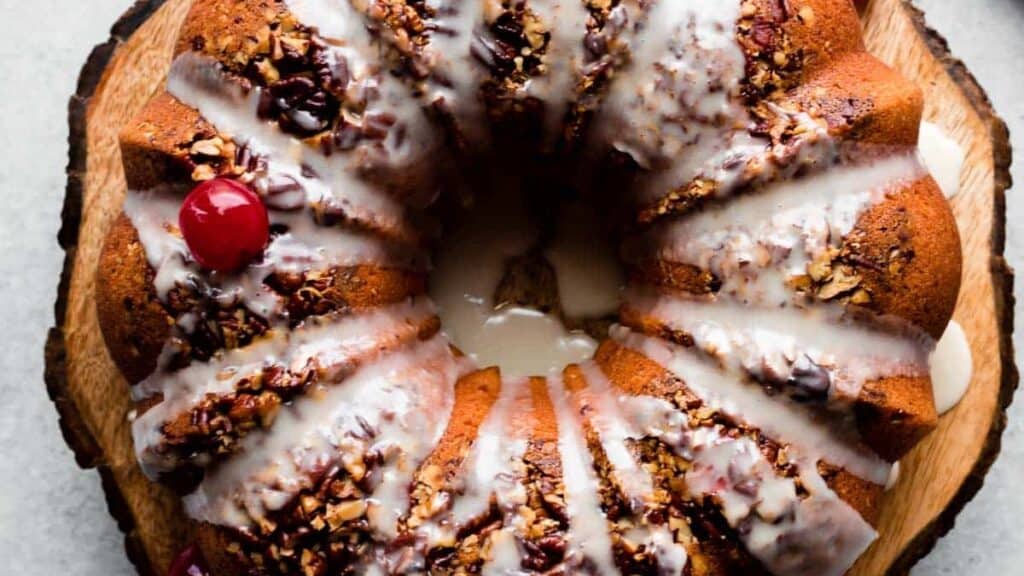 A bundt cake with icing drizzled on top, garnished with glazed cherries and chopped nuts. It is placed on a wooden serving board.