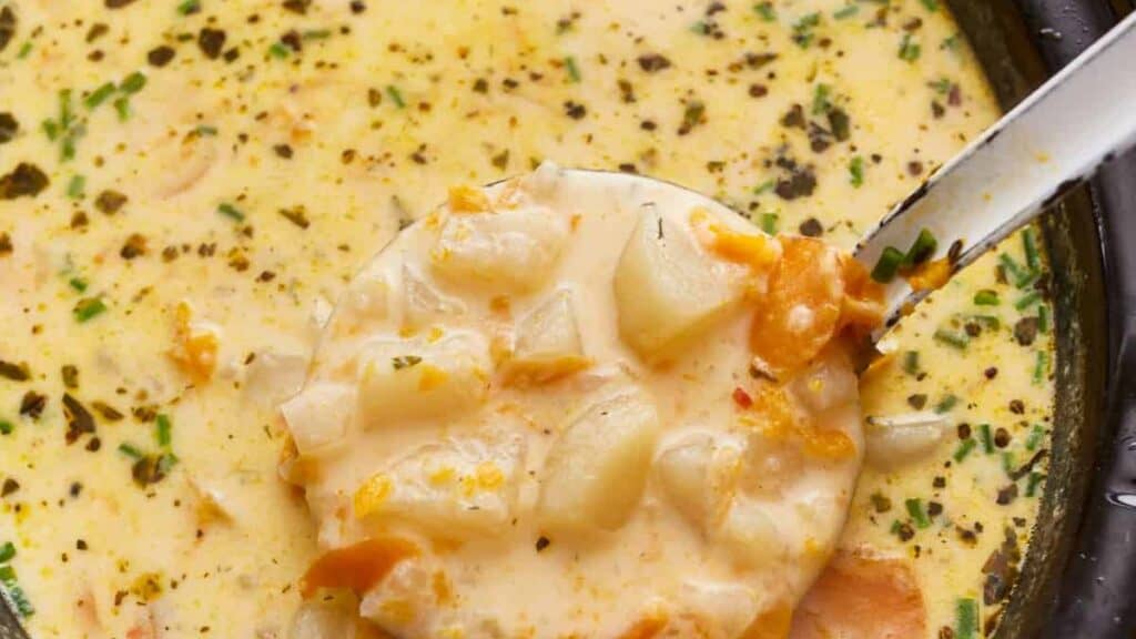 A close-up of a creamy soup with chunks of potato and carrots, garnished with herbs. A ladle is lifting a portion from the pot, showing the rich, textured consistency.