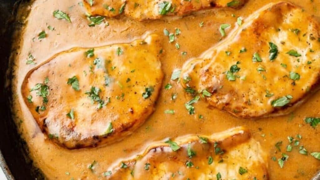 Four golden-brown pork chops simmer in a rich, creamy brown sauce, garnished with chopped parsley in a pan.