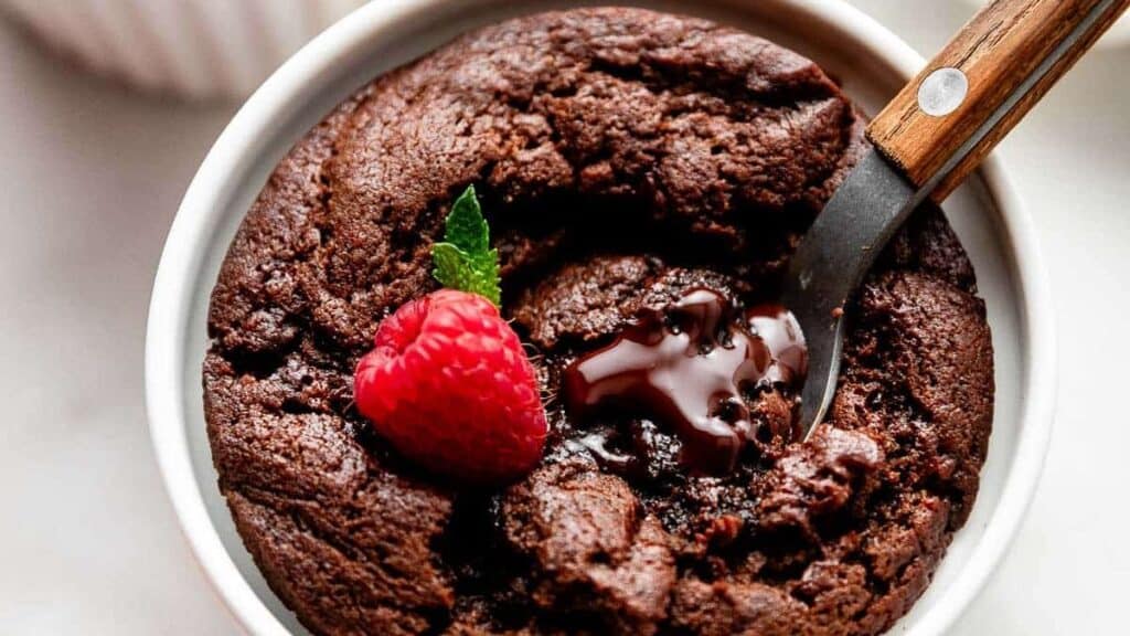 A rich chocolate lava cake is served in a white ramekin, with melted chocolate oozing from the center. It is garnished with a fresh raspberry and a small mint leaf, and a wooden-handled spoon rests on the edge.