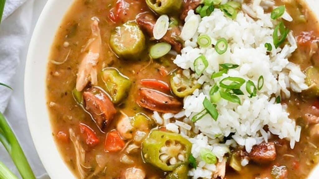 A bowl of gumbo with sliced sausage, chicken, okra, and diced red peppers in a thick broth. It's topped with a serving of white rice and garnished with chopped green onions.