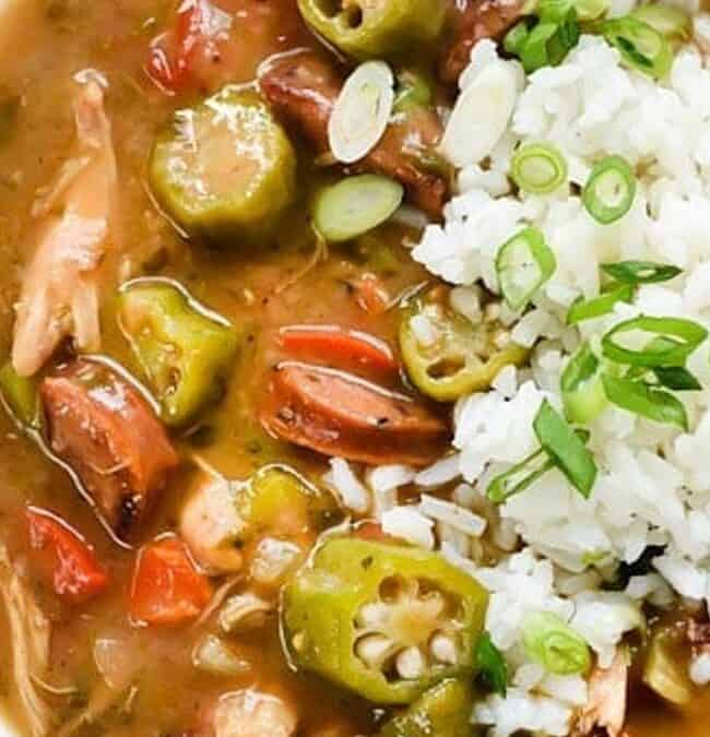 A bowl of gumbo with sliced sausage, chicken, okra, and diced red peppers in a thick broth. It's topped with a serving of white rice and garnished with chopped green onions.