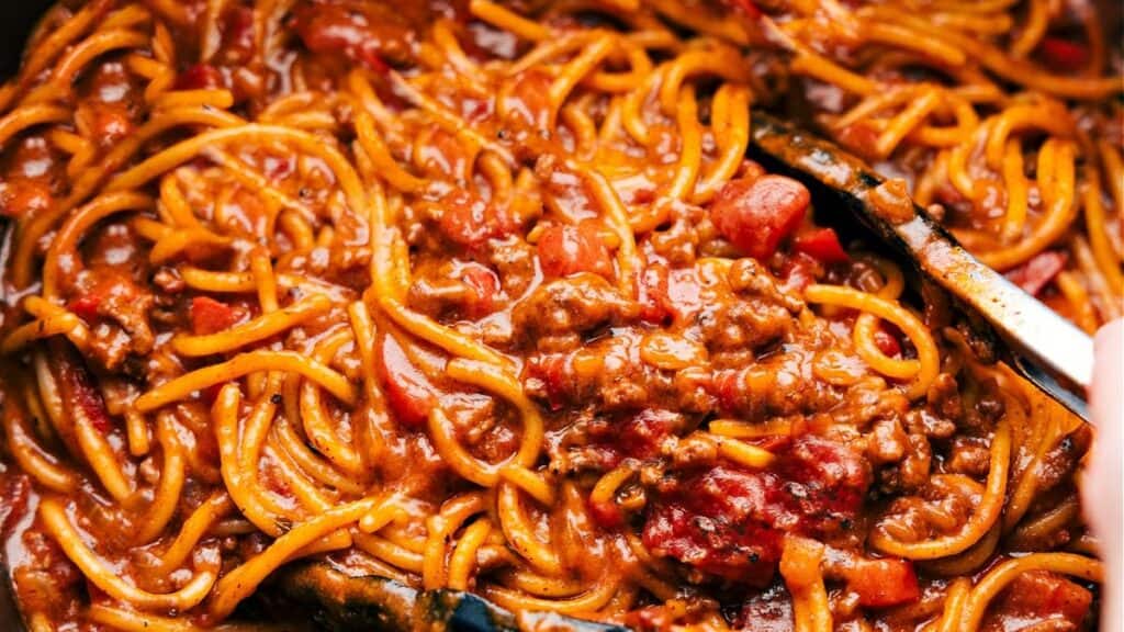 Close-up of spaghetti mixed with a rich, meaty tomato sauce. This comfort food features pasta coated in a thick sauce with chunks of tomatoes and ground beef, crafting a hearty and savory dish. A serving utensil is partially visible in the corner.