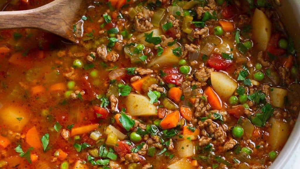 A hearty vegetable soup with ground meat, green peas, chopped carrots, diced potatoes, and tomatoes. Garnished with fresh parsley, the soup is in a pot with a wooden spoon, showcasing a vibrant mix of colors and textures.