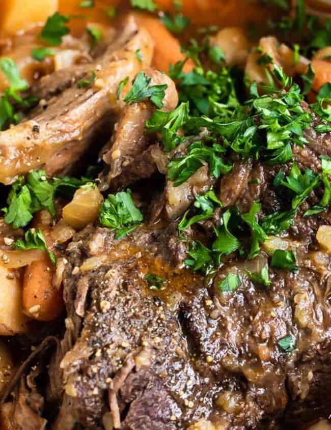 A pot filled with a classic, old-fashioned pot roast surrounded by carrots, potatoes, and onions. The dish is garnished with chopped parsley, and there is a rich, savory sauce visible in the pot.