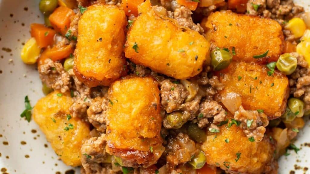 A close-up of a savory dish featuring crispy tater tots on top of a flavorful mixture of ground meat, peas, carrots, corn, and onions, garnished with parsley. The dish is served on a white speckled plate.