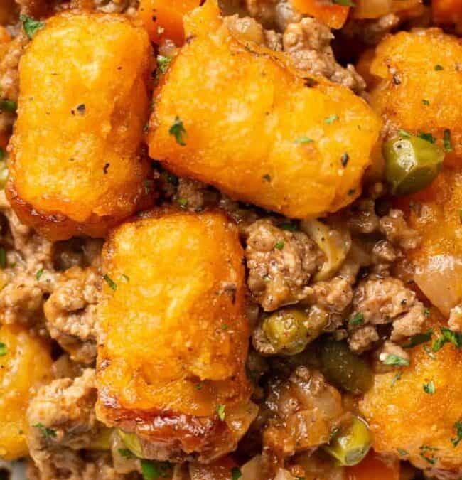 A close-up of a savory dish featuring crispy tater tots on top of a flavorful mixture of ground meat, peas, carrots, corn, and onions, garnished with parsley. The dish is served on a white speckled plate.