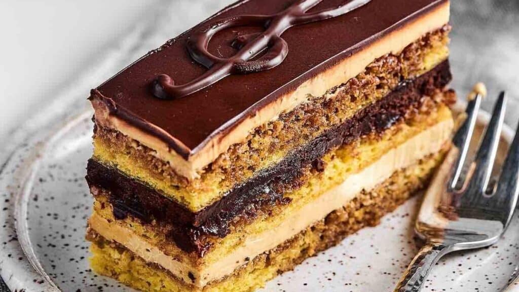 A slice of layered opera cake on a speckled plate captures the essence of timeless desserts. The cake showcases thin sponge layers, coffee cream, and chocolate ganache beneath a glossy glaze, reminiscent of 1960s retro desserts. A fork rests beside the enticing slice.