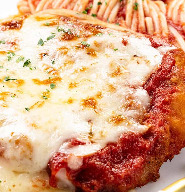 Close-up of chicken parmesan topped with melted cheese and sauce, accompanied by a side of spaghetti. The dish is garnished with herbs, and a fork rests nearby on a white plate.