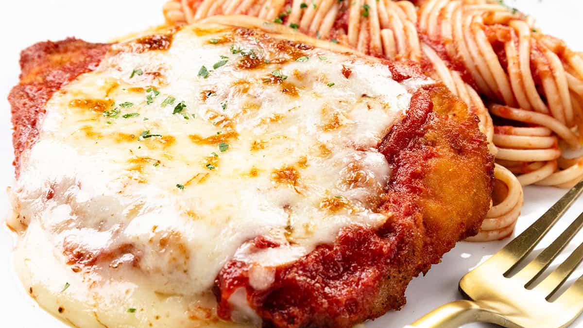 Close-up of chicken parmesan topped with melted cheese and sauce, accompanied by a side of spaghetti. The dish is garnished with herbs, and a fork rests nearby on a white plate.