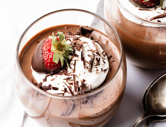 Two glass cups filled with chocolate mousse are topped with whipped cream, showcasing delightful ways to use whipped cream. Each is adorned with a fresh strawberry and chocolate shavings, while a silver spoon rests elegantly beside them on the white surface.