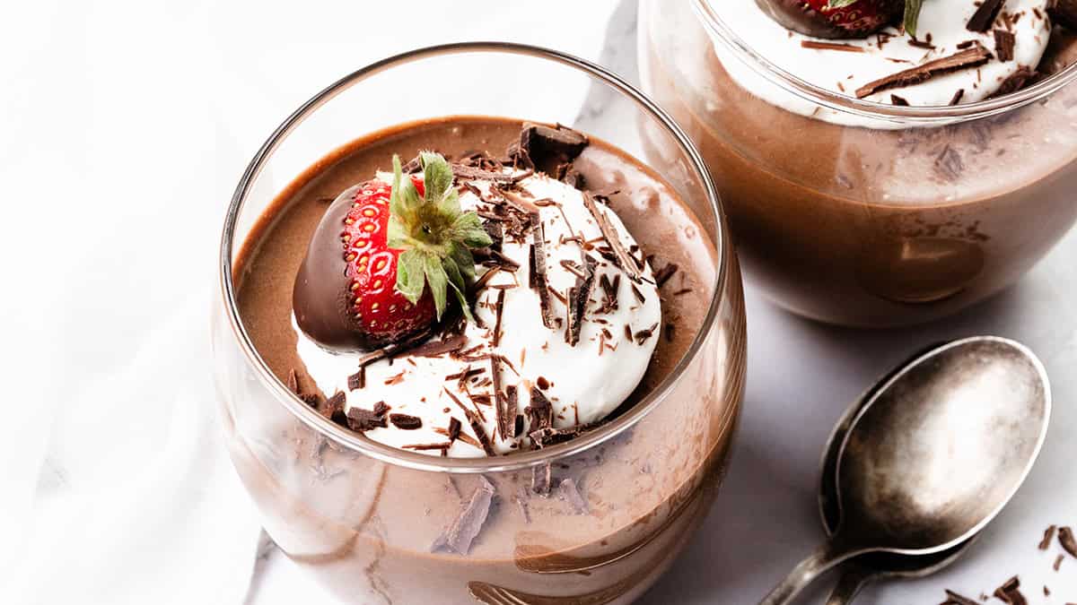 Two glass cups filled with chocolate mousse are topped with whipped cream, showcasing delightful ways to use whipped cream. Each is adorned with a fresh strawberry and chocolate shavings, while a silver spoon rests elegantly beside them on the white surface.