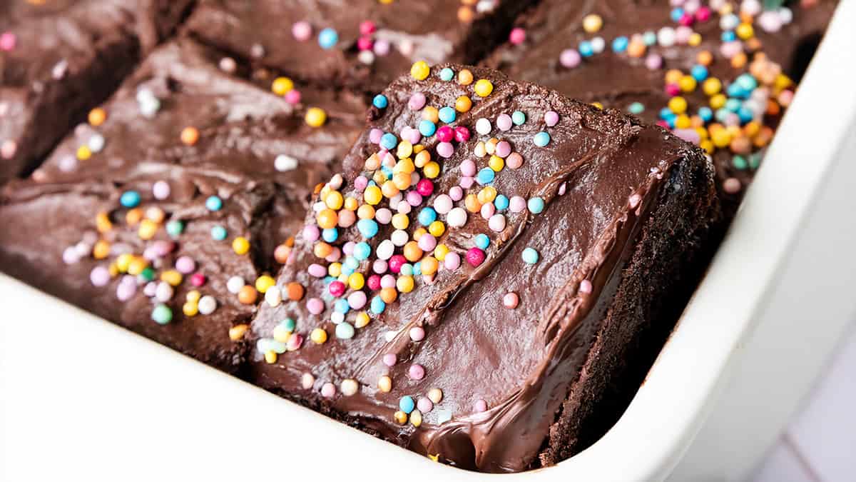 A close-up of chocolate cake pieces in a white dish. One piece is tilted, showing a rich, glossy chocolate frosting topped with colorful round sprinkles.