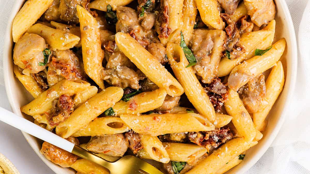 A bowl of creamy penne pasta with pieces of chicken, sun-dried tomatoes, and fresh basil, topped with a sprinkle of grated cheese. A gold fork rests inside the bowl, ready for eating.