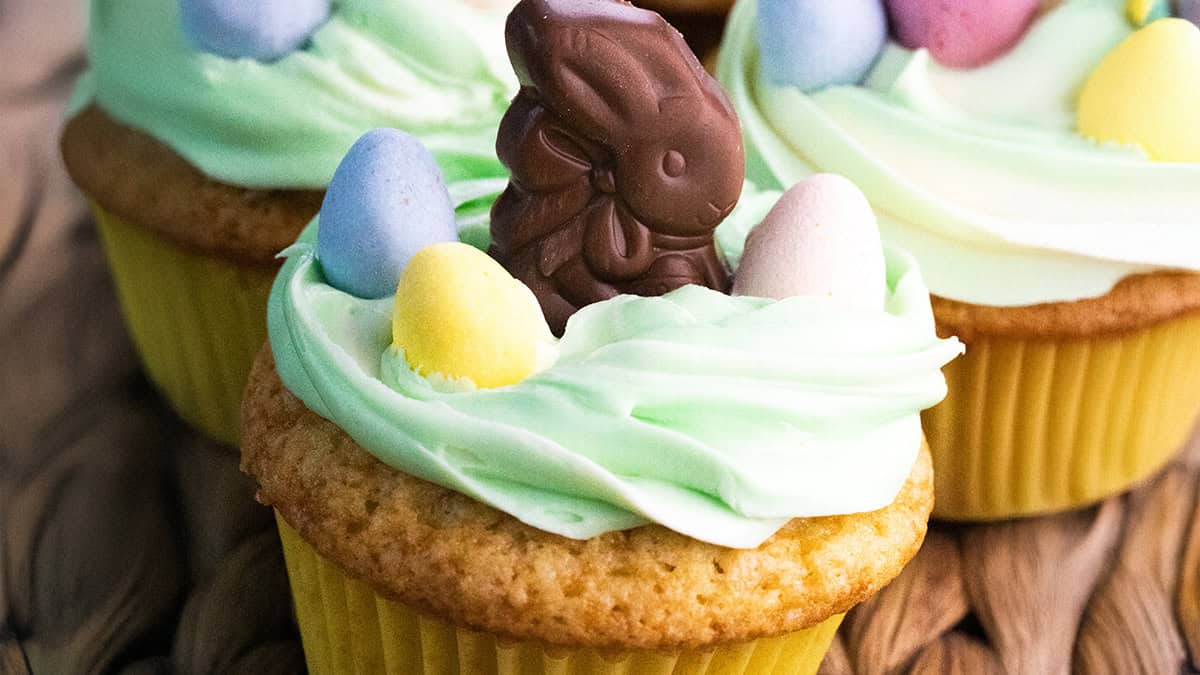 Cupcake with light green frosting topped with a chocolate bunny and colorful candy eggs. It sits in a yellow paper liner, alongside similar decorated cupcakes.