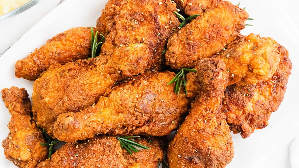 A plate filled with crispy fried chicken drumsticks and wings, garnished with sprigs of fresh rosemary. The chicken is golden-brown and seasoned with herbs, set on a white platter.