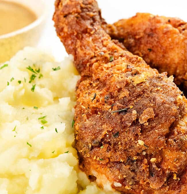 Close-up of crispy fried chicken drumsticks with a side of creamy mashed potatoes sprinkled with herbs. A small bowl of brown gravy is in the background on a white plate.