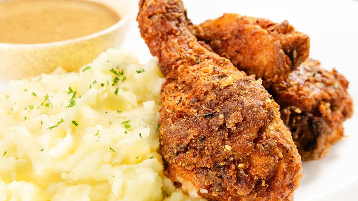 Close-up of crispy fried chicken drumsticks with a side of creamy mashed potatoes sprinkled with herbs. A small bowl of brown gravy is in the background on a white plate.