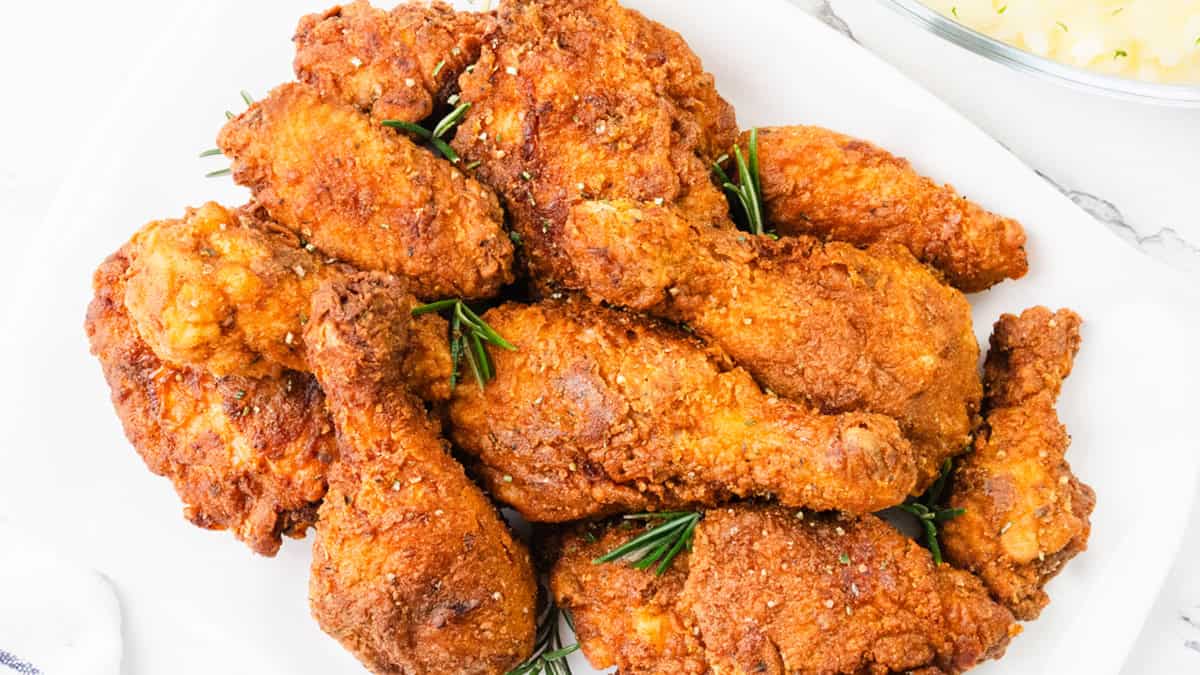 A plate of golden-brown fried chicken pieces, garnished with sprigs of rosemary, placed on a white rectangular platter. The chicken looks crispy and seasoned, with a glass bowl partially visible in the background.