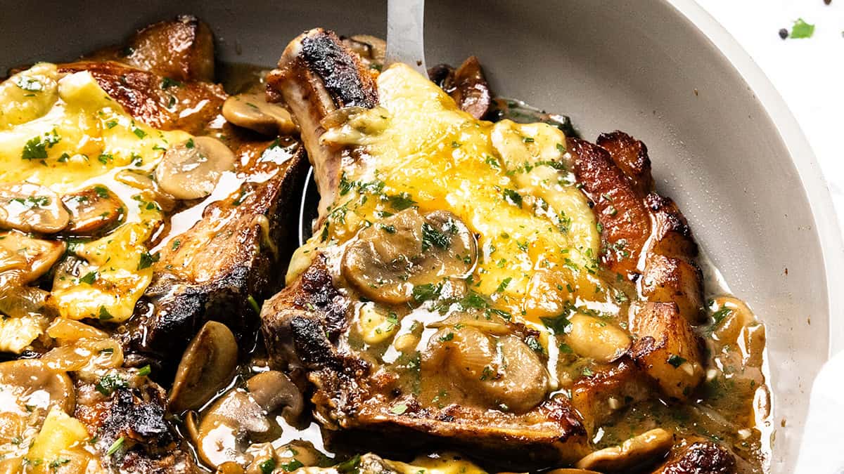 A close-up of pork chops in a skillet, smothered with creamy mushroom sauce and melted cheese. Mushrooms and herbs crown the dish, giving it a slightly browned and savory look—perfect for those exploring new pork chop recipes.