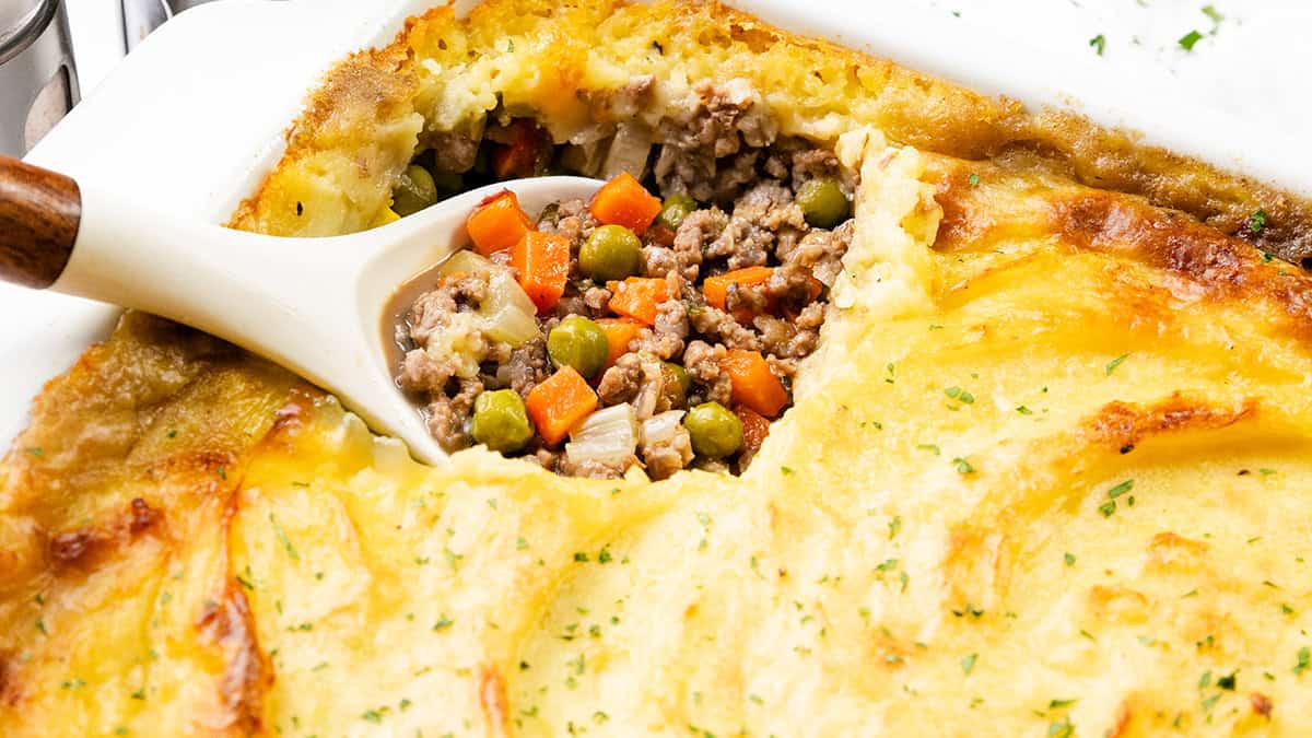 A baked shepherd's pie with a golden, crispy mashed potato topping is shown with a portion removed, revealing a filling of ground meat, carrots, peas, and onions in a brown sauce. A serving spoon is scooping out the filling.