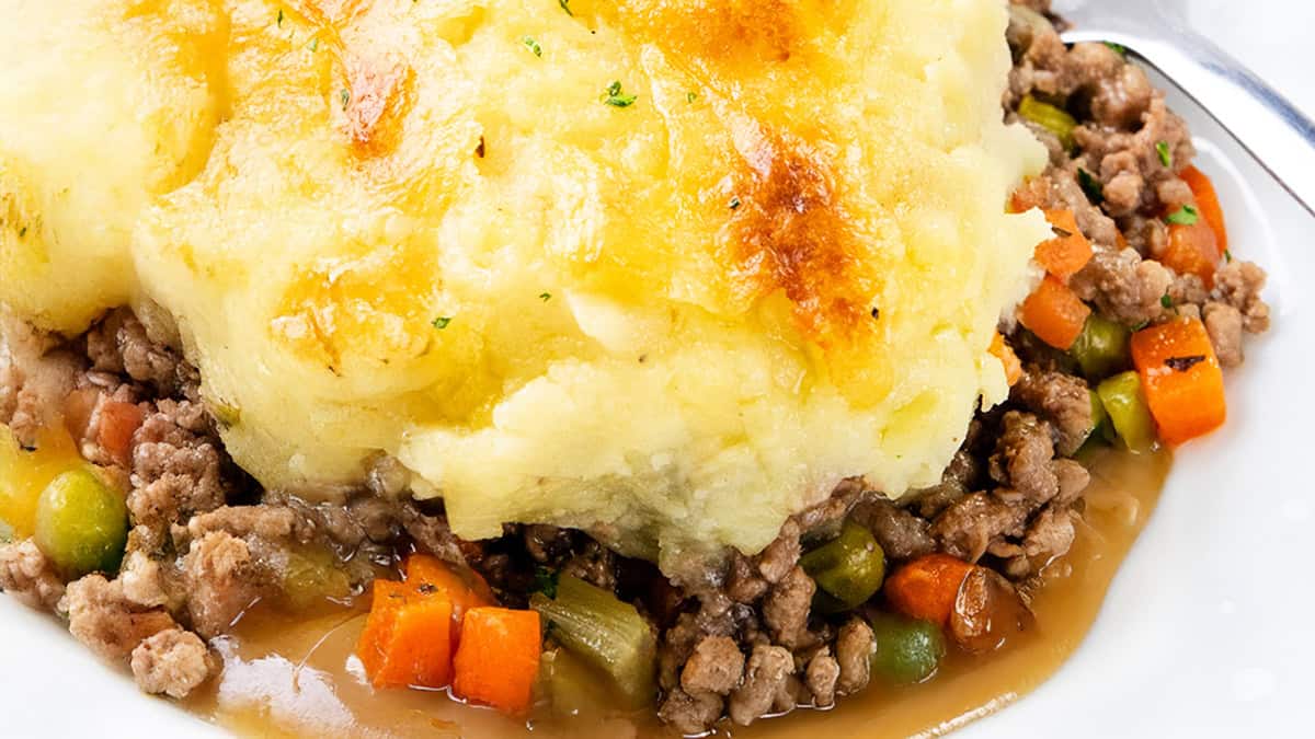 Close-up of a shepherd's pie featuring a golden-brown mashed potato topping. Below, there's a mixture of cooked ground meat, peas, carrots, and other vegetables, all in a brown gravy, served on a white plate.