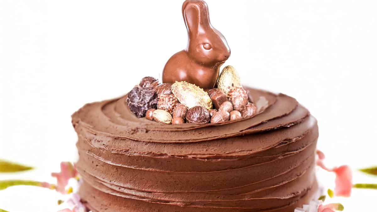 A chocolate cake with smooth frosting is topped with a chocolate bunny and assorted chocolates. The cake sits on a white surface with blurred pink flowers around the base.