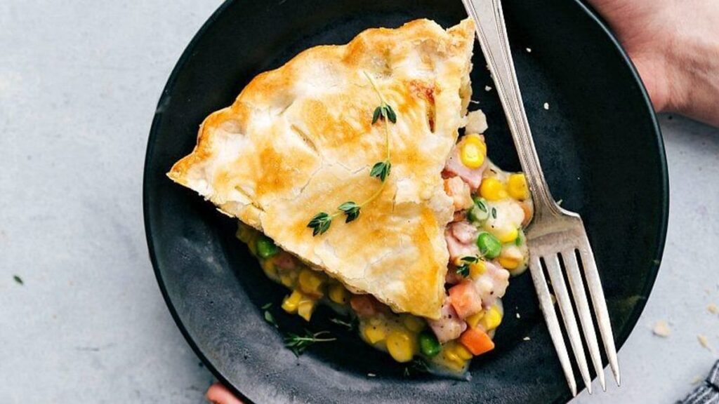 A slice of vegetable pot pie on a black plate, garnished with fresh herbs. The pie has a golden, flaky crust and is filled with peas, corn, and carrots. A fork rests beside the slice.